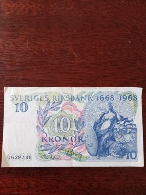 Svensk 10-kronorssedel 1668-1968 - Unik 10-kronorssedel från Sveriges Riksbank, utgiven 1968 för att fira 300-årsjubileet. Sedeln har detaljerade motiv i blått, grönt och rosa samt texten 'SVERIGES RIKSBANK 1668-1968'. Baksidan visar en byggnad och folkmassa i rött.