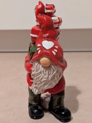 Tomte med hjärtdetalj - Unik tomte i rött med vit hjärtdetalj på luvan, gröna inslag och svarta skor. Tomten har en klassisk julstil och är dekorerad med flera paket i rött, vitt och grönt. Perfekt för dig som vill ha en färgglad tomte som dekoration.