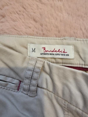 Beige chinos från Bondelid M - Snygga beige chinos från Bondelid i storlek M. Klassisk rak passform med bälteshällor, knappgylf och diskreta fickor. Detaljer i rött vid fickorna och insidan av linningen ger en cool touch. Perfekta för en avslappnad men stilren look.