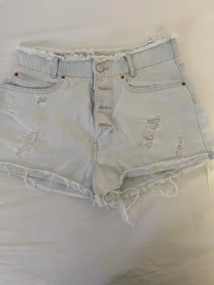 Ljusa denimshorts från Pull&Bear - Säljer ett par ljusa denimshorts från Pull&Bear med hög midja, slitna detaljer och fransig kant. Shortsen har knappar framtill, klassiska fickor och en avslappnad passform. Perfekta för varma dagar och en chill stil.