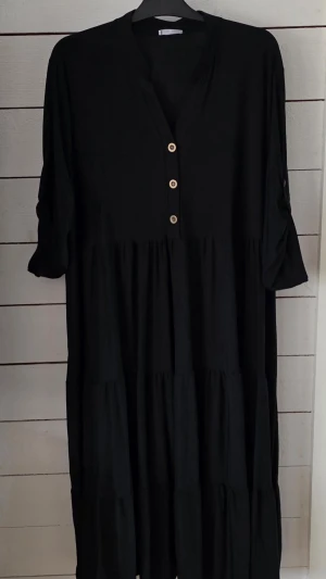 Svart långklänning från Zanca Sonne - Svart långklänning från Zanca Sonne med v-ringning och tre dekorativa knappar framtill. Klänningen har trekvartsärm och ett ledigt, flowy fall. Perfekt för dig som gillar enkel och stilren design.