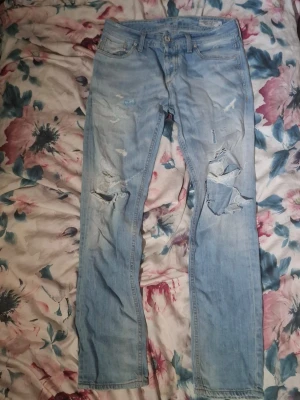 Ljusblå slitna jeans med hål - Säljer ett par ljusblå jeans med slitningar och stora hål på knäna. Jeansen har klassisk femficksdesign och raka ben. Materialet är denim och de har en cool, avslappnad look som passar perfekt till sneakers.