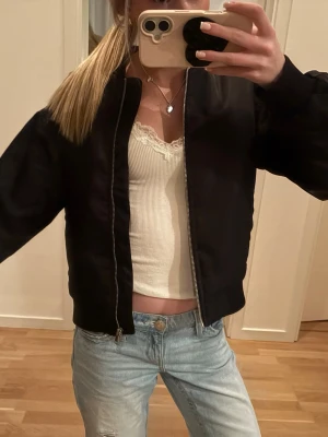 Svart bomberjacka med dragkedja - Stilren svart bomberjacka med klassisk passform och silverfärgad dragkedja framtill. Jackan har ribbade muddar vid ärmslut och midja. Perfekt att slänga över en topp och jeans för en cool look.