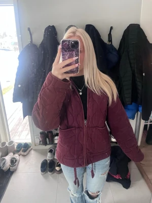 Vinröd vårjacka - Säljer en vinröd vårjacka med hög krage och dragkedja framtill. Jackan har ett vågigt mönster i tyget, två stora fickor och justerbar dragsko. Se bilder på små skavanker!