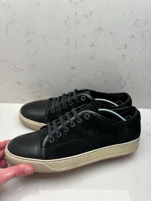 Lanvin skor  - Stilrena svarta sneakers med ovandel i mocka och tåparti i slätt läder. Skorna har svarta skosnören, metallöljetter och en kontrasterande vit sula. Perfekta för en clean och avslappnad look.