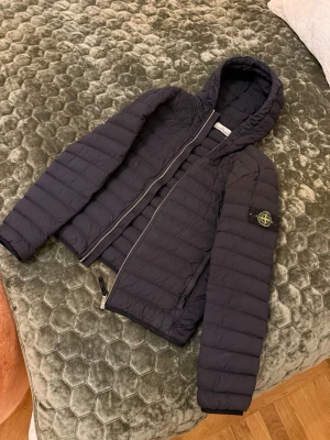 Svart lättviktsjacka Stone Island - Säljer en svart lättviktsjacka från Stone Island med huva och quiltad design. Jackan har dragkedja framtill och den klassiska Stone Island-loggan på vänster ärm. Perfekt för dig som vill ha en stilren och sportig look. Kvittot har jag tyvärr inte kvar. Men den är inköpt på Stone Island i Stockholm. Och det går att verifiera att den är äkta. 
