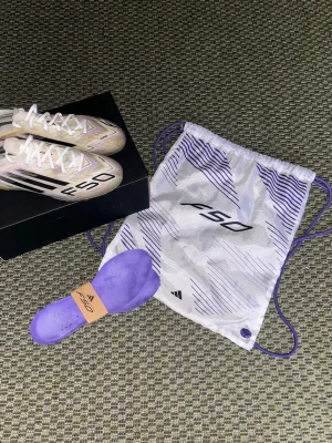 Adidas F50 fotbollsskor gul/lila elites - Säljer ett par Adidas F50 fotbollsskor i gul och lila med svarta detaljer och vita skosnören. Skorna har en lätt och aerodynamisk form, AG-dobbar för spel på konstgräs och kommer med en matchande påse samt extra innersula. Perfekta för dig som vill ha speed och stil på planen.