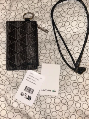 Svart monogram plånbok från Lacoste - Snygg svart plånbok från Lacoste med geometriskt monogrammönster och silverfärgad nyckelring. Plånboken är tillverkad i polyvinylklorid och har ett praktiskt snöre för att hänga runt halsen. Perfekt för att hålla koll på kort och nycklar.