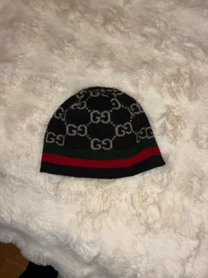 Svart Gucci mössa med logga - Säljer en svart stickad mössa från Gucci med klassiskt GG-mönster i vitt och gröna samt röda ränder längst ner. Mössan har en tight passform och är tillverkad i mjukt material som känns skönt mot huden. Perfekt accessoar för dig som gillar exklusiv streetwear.