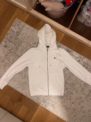 Vit hoodie från Polo Ralph Lauren - Snygg vit hoodie från Polo Ralph Lauren med dragkedja framtill och klassisk broderad logga på bröstet. Hoodien har två fickor fram, ribbade muddar och justerbar huva med snören. Perfekt för en clean och stilren look.