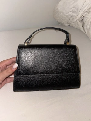 Svart stilren handväska i skinnimitation - Snygg svart handväska i skinnimitation med enkel, stilren design och guldfärgade detaljer vid handtaget. Har använt ett par gånger, det tillkommer inget band med eftersom att den är borta. Annars är det en väldigt fin väska