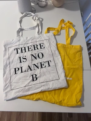 Två tygkassar med tryck - Säljer två shoppingväskor i tyg, en vit med svart text 'THERE IS NO PLANET B' och en gul med vit text 'Sjukt bra i Kristianstads kommun'. Båda har långa handtag och är perfekta för att bära böcker, mat eller annat du behöver. Materialet är bomull och de är lätta att vika ihop.