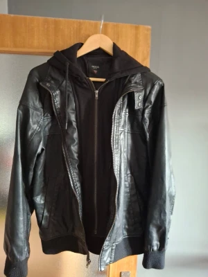 Svart skinnjacka från Guess - Snygg svart skinnjacka från Guess med dragkedja, ribbade muddar och huva. Jackan har coola detaljer som tryckknappar vid axlarna och flera fickor. Perfekt för dig som vill ha en stilren men ändå edgy look. Lite slitage på vänster arm, se bild 