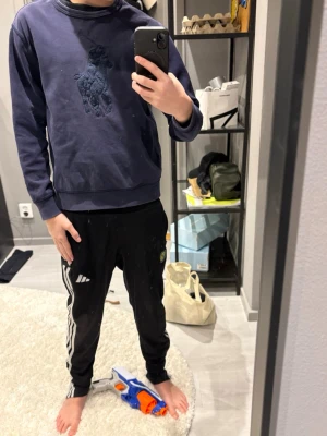 Mörkblå sweatshirt från Polo Ralph Lauren - Mörkblå sweatshirt från Polo Ralph Lauren med stor ton-i-ton broderad logga på bröstet. Klassisk rund halsringning och ribbade muddar vid ärmslut och nederkant. Perfekt för en avslappnad och stilren look.