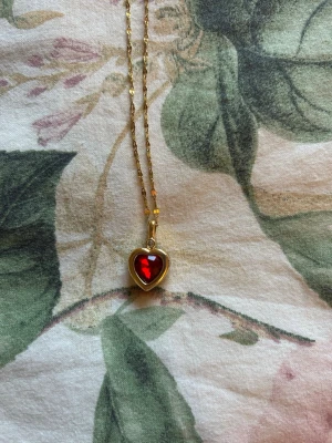 Guldhalsband med rött hjärthänge - Snyggt halsband i guldfärgad metall med ett hjärtformat hänge. Hänget har en röd, glittrande sten i mitten som ger en romantisk vibe. Kedjan är tunn och elegant, perfekt för att lyfta vilken outfit som helst. 