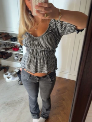 Svartvitrutig blus med volang - Söt svartvitrutig blus med korta puffärmar och volang nedtill. Toppen har en markerad midja och v-ringad urringning som ger en feminin siluett. Perfekt till jeans eller kjol för en trendig look.