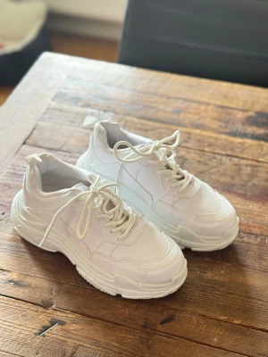 Chunky vita sneakers med tjock sula - Stilrena chunky sneakers i helvitt med tjock, mönstrad sula och klassisk snörning. Skorna har en modern, sportig look och är tillverkade i syntetmaterial med textildetaljer. Perfekta för dig som gillar trendiga och bekväma sneakers med extra höjd.