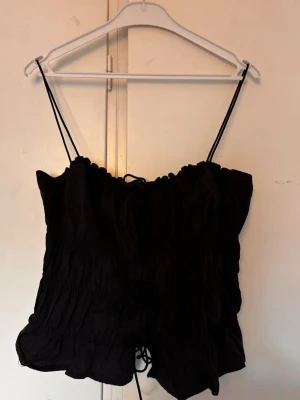 Svart linnetopp  - Ej använd svart linnetopp med tunna axelband och snörning framtill. Toppen har en rynkad design och volangkant nedtill som ger en söt och trendig look. Perfekt att matcha med jeans eller kjol för en chill stil.