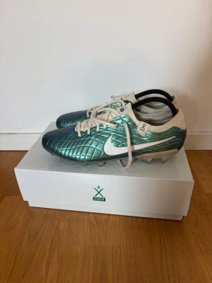 Nike fotbollsskor turkos/vit 2024 - Snygga fotbollsskor från Nike i metallic turkos med vita detaljer och snörning. Skorna har quiltad ovandel och är designade för gräsplaner. Lätta och smidiga med modern look, perfekta för dig som vill sticka ut på planen. Levereras i originalkartong med 2024-märkning.