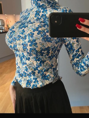 Blommig polobody i blått och vitt - Säljer en slim polotopp med blått blommönster på vit botten. Toppen har lång ärm och hög krage, sitter tight och är perfekt för dig som gillar färgglada prints. Materialet känns stretchigt och mjukt, troligen polyester. Superfin till både jeans och kjol! Aldrig använd. 