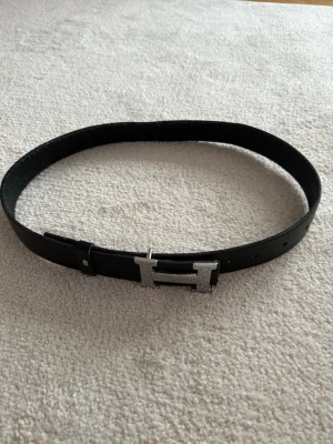 Svart skärp med H-spänne - Snyggt svart bälte med ett stort H-format spänne i silverfärg. Bältet har en stilren och klassisk look som passar till många outfits. Tillverkat i skinn för en exklusiv känsla och lång hållbarhet. Köparen står för frakt om den ska fraktas!