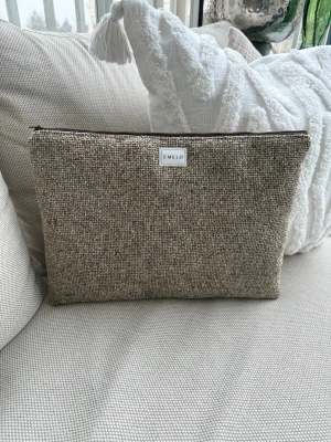 Beige kuvertväska från Emelo - Stilren datorväska från Emelo i beige och brunt vävt tyg. Väskan har en rektangulär form, dragkedja upptill och en ljus insida. Perfekt för att bära din laptop med en snygg och enkel look. Passar laptop med maxmått: 33x24cm
