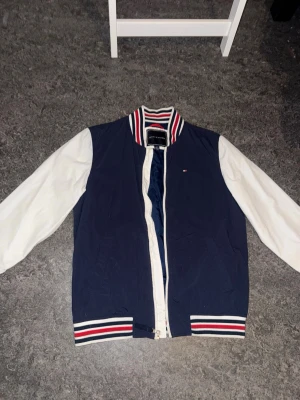 Blåvit bomberjacka Tommy Hilfiger - Snygg bomberjacka från Tommy Hilfiger i marinblått med vita ärmar. Jackan har ribbade muddar och krage med röd, vit och blå randig detalj samt dragkedja framtill. Liten logga på bröstet och klassisk college-look. Perfekt för dig som gillar streetwear och sportig stil.