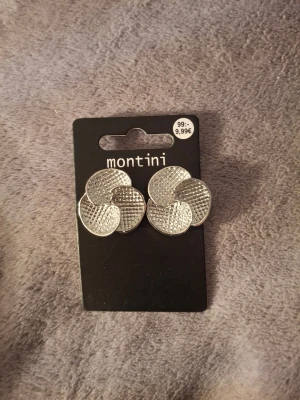 Silvriga örhängen från Montini - Snygga silvriga örhängen från Montini med rund, snurrad design och strukturerad yta som ger ett coolt intryck. Perfekta för att lyfta vilken outfit som helst med sin unika form och metalliska finish.