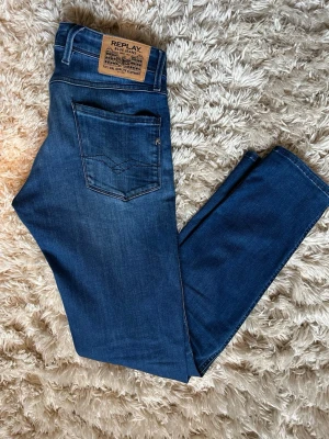 Blåa Replay Anbass - Snygga blå jeans från Replay med klassisk femficksmodell och raka ben. Jeansen har en mörk tvätt med lätt slitning framtill och detaljer i silverfärg. Tillverkade i bomull med en skön stretch för extra komfort. Perfekta för en avslappnad och trendig look.