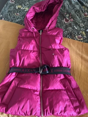 Rosa dunväst från Armani Exchange - Snygg rosa dunväst från Armani Exchange med huva och dragkedja framtill. Västen har ett markerat midjebälte med logga och två stora fickor. Perfekt för lager på lager och ger en cool streetstyle-look.