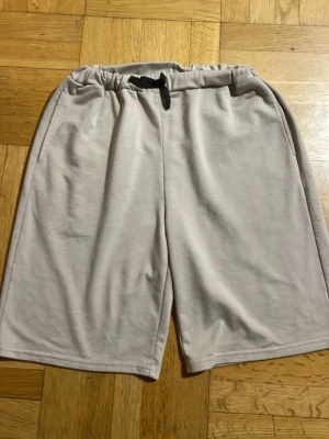 Ljusgrå mjuka shorts med snörning - Säljer ett par beige shorts med elastisk midja och svart snörning. Shortsen har en loose passform och är tillverkade i ett mjukt syntetmaterial, perfekta för avslappnade dagar. Två sidofickor för extra bekvämlighet.