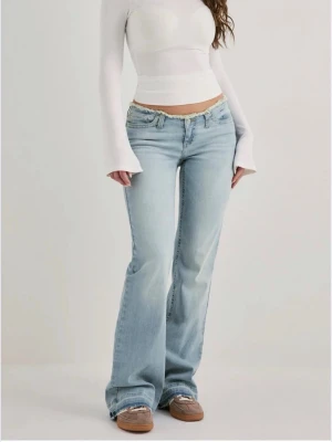 Ljusblå bootcut jeans med låg midja - Säljer ett par ljusblå bootcut jeans med låg midja och slitna detaljer vid linningen. Jeansen har klassisk femficksdesign och är tillverkade i denim med lätt tvättad look. Perfekta för en avslappnad och trendig stil. Från Nelly använda en gång storlek 36/short.