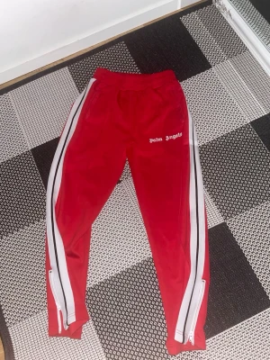Röda Palm Angels mjukisbyxor - Säljer ett par röda mjukisbyxor från Palm Angels med vita och svarta sidoremsor. Byxorna har resår i midjan, dragkedjor vid bensluten och logga broderad på vänster sida. Perfekta för en chill och sportig look.