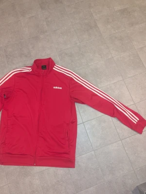 Röd Adidas track jacket - Snygg röd Adidas track jacket med klassiska vita ränder längs ärmarna och vit Adidas-logga på bröstet. Jackan har dragkedja framtill, hög krage och två sidofickor. Perfekt för träning eller streetwear. Tillverkad i ett mjukt och stretchigt material.