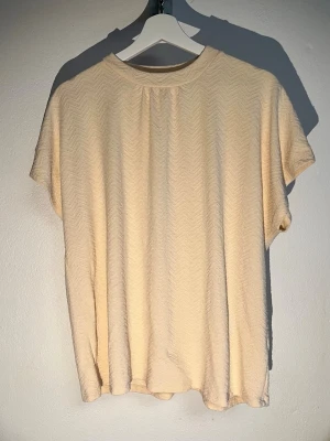 Lindex cremefärad stopp med strukturmönster storlek L - Säljer en beige t-shirt med diskret zigzag-mönster i tyget. T-shirten har korta ärmar och en avslappnad passform. Materialet känns mjukt och luftigt, perfekt för varmare dagar. Enkel och stilren design som passar till det mesta.