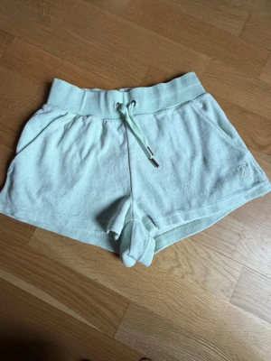Ljusgröna shorts från Juicy Couture - Säljer ett par ljusgröna shorts från Juicy Couture  med bred resår i midjan och snörning framtill. Shortsen har fickor på sidorna och en liten broderad logga på ena benet. Perfekta för sommardagar eller chill hemma.