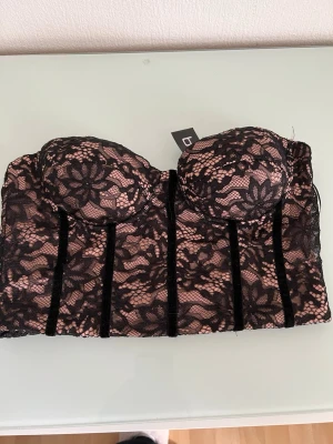 Svart spetsbustier från Boohoo - Snygg bustier från Boohoo i svart spets med blommönster och nude-färgad bas. Kuporna är vadderade och modellen har markerade sömmar framtill för extra form. Dragkedja bak för enkel påklädning. Perfekt att styla under en kavaj eller transparent topp.