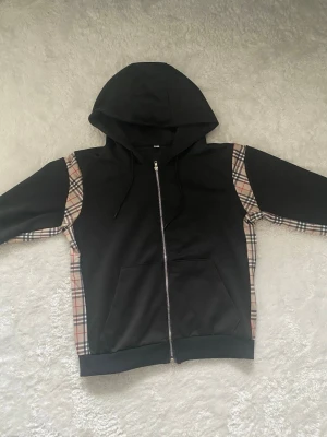 Svart hoodie med rutiga detaljer - Snygg svart hoodie med dragkedja och klassiska beige-rutiga paneler längs ärmar och sidor. Utrustad med stor huva och känguruficka framtill. Perfekt för dig som gillar streetwear och vill sticka ut lite extra. Materialet känns mjukt och bekvämt.