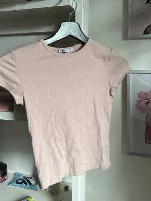 Beige t-shirt från Bershka - En enkel och stilren beige t-shirt från Bershka. Modellen är klassisk med rund halsringning och korta ärmar. Tillverkad i mjuk bomull som känns skön mot huden. Perfekt basplagg att matcha med allt i garderoben. Sitter tight.
