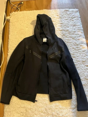 Svart hoodie från Nike med dragkedja - Snygg svart hoodie från Nike med dragkedja framtill och huva. Hoodien har diskreta svarta detaljer över bröstet och två fickor på sidorna. Tillverkad i mjukt material som passar perfekt till en avslappnad streetwear-look.