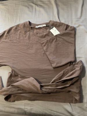 Brun t-shirt som man knyter bak från Lager 157 - En enkel brun t-shirt som man knyter bak från Lager157 med rund hals och korta ärmar. Tillverkad i mjuk bomull som känns skön mot huden. Storlek S. Den är helt ny och oanvänd