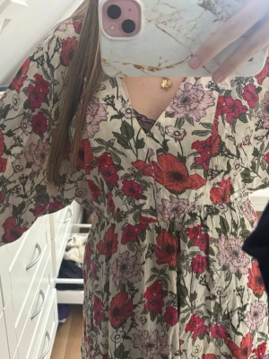 Blommig omlottklänning med v-ringning - Säljer en luftig blommig klänning med v-ringning och omlottkänsla. Klänningen har korta ärmar, resår i midjan och ett mönster i rött, rosa och grönt på beige botten. Perfekt för varma dagar och ger en romantisk vibe.