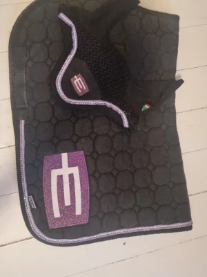 Svart schabrak och huva med lila detaljer - Säljer ett svart schabrak och matchande huva från Equiline med lila och vita detaljer. Båda har ett stort lila logomärke och kantband i lila. Schabraket har bikakemönster och huvan är virkad med elastiska öron.  strl ponny