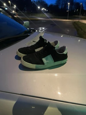 Svarta sneakers från Valentino - Snygga svarta sneakers från Valentino med vita detaljer och en bred vit rem på sidan. Skorna har en låg silhuett och klassisk snörning. Yttermaterialet ser ut att vara mocka eller läder och sulan är vit med nitar baktill. Perfekta för en trendig streetwear-look.