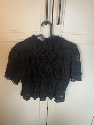 Svart spetsblus med volangdetaljer - Trendig svart blus med korta ärmar och volangdetaljer. Blusen är gjord i spets och har en lätt croppad passform. Perfekt för dig som gillar romantiska detaljer och vill ha en unik topp i garderoben.