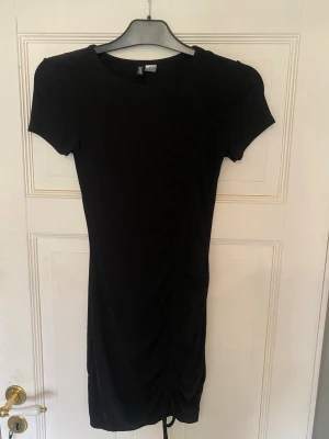 Svart kort klänning från H&M - En svart, kort klänning från H&M med rund halsringning och korta ärmar. Klänningen har en enkel, figurnära passform och snygg rynkdetalj med snörning på sidan. Perfekt att styla med sneakers eller boots.