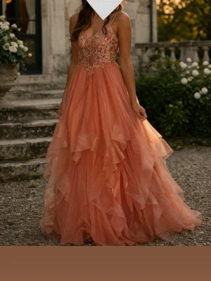 Rosa långklänning med spetsdetaljer - Romantisk rosa långklänning med tunna axelband och vacker spets upptill. Klänningen har volangkjol i flera lager och snörning i ryggen för en justerbar passform. Perfekt för speciella tillfällen där du vill känna dig extra fin.