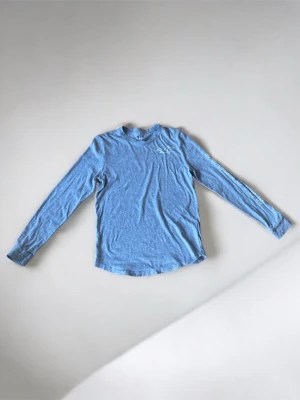 Blå långärmad topp från Hollister - Ljusblå långärmad topp från Hollister med rund halsringning och tryckt logga på bröstet samt större logga på ryggen. Toppen har även tryck längs ena ärmen och är tillverkad i mjuk bomullsmix för en bekväm känsla.