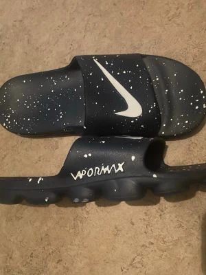 Nike Vapormax svarta sandaler - Svarta Nike Vapormax sandaler med vit stänkmönster och stor vit Swoosh-logga på remmen. Sandalerna har en chunky sula och öppen tå, tillverkade i syntetmaterial. Perfekta för en avslappnad och sportig stil.