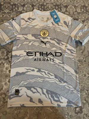 Manchester City bortatröja Puma - Snygg Manchester City bortatröja från Puma i vitt och ljusgrått med drakmönster. Tröjan har klubbmärke på bröstet, svart Puma-logga och Etihad Airways-tryck. Tillverkad i lätt och ventilerande funktionsmaterial, perfekt för fotboll eller träning.